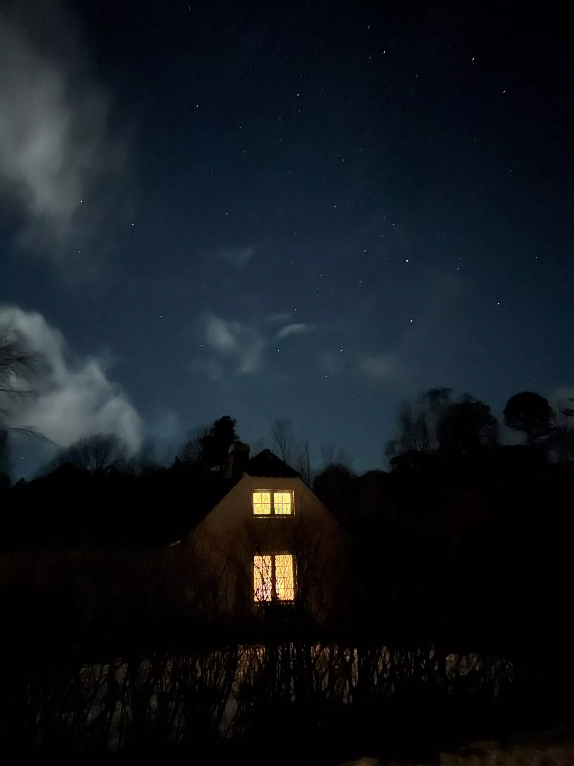 Et lille hus med lysende gule vinduer står i mørket, omgivet af træer og buske under en stjerneklar nattehimmel med spredte skyer.