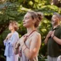 En gruppe mennesker står udendørs med lukkede øjne og hænderne på brystet og ser rolige og meditative ud, omgivet af frodigt grønt løv.