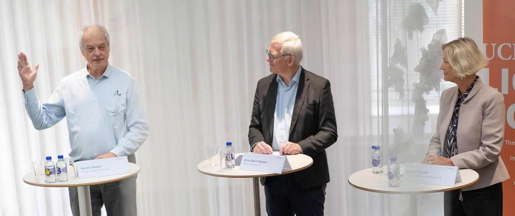 Tre personer står ved runde borde og diskuterer. Manden til venstre gestikulerer med løftet hånd, mens den anden mand og kvinde lytter opmærksomt. Navneskilte og vandflasker står på bordene, og i baggrunden er der hvide gardiner.