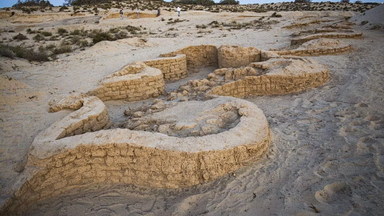 Gamle, delvist begravede murstensruiner i et sandet ørkenlandskab med sparsom vegetation. Et par mennesker ses i det fjerne, og arabisk tekst vises i nederste højre hjørne.