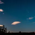 En nattehimmel med spredte skyer, stjerner og en komet med en synlig hale. I bunden ses silhuetter af træer og et par huse, og svage flylys strejfer hen over himlen.