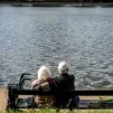 Et ældre par sidder på en bænk ved en sø med ryggen til kameraet. Den ene person har en arm om den anden. En rollator og en paraply står ved siden af bænken, og vandet fylder baggrunden.