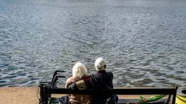 Et ældre par sidder på en bænk ved en sø med ryggen til kameraet. Den ene person har en arm om den anden. En rollator og en paraply står ved siden af bænken, og vandet fylder baggrunden.