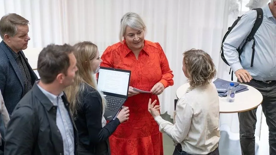 En gruppe på fem voksne har en diskussion omkring et højt rundt bord. En kvinde i rød kjole står i midten, mens en anden kvinde holder en bærbar computer, og andre lytter eller taler. De befinder sig indendørs med hvide gardiner i baggrunden.