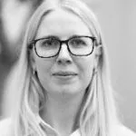 En kvinde med langt, glat blondt hår og briller med mørkt stel ser direkte ind i kameraet. Hun er iført en lys top, og baggrunden er sløret. Billedet er i sort-hvid.