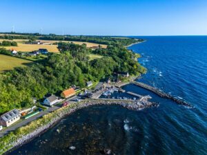 Luftfoto af en kystby med farverige huse, en lille havn, grønne marker, træer og vindmøller i baggrunden ved et roligt blåt hav under en klar himmel.