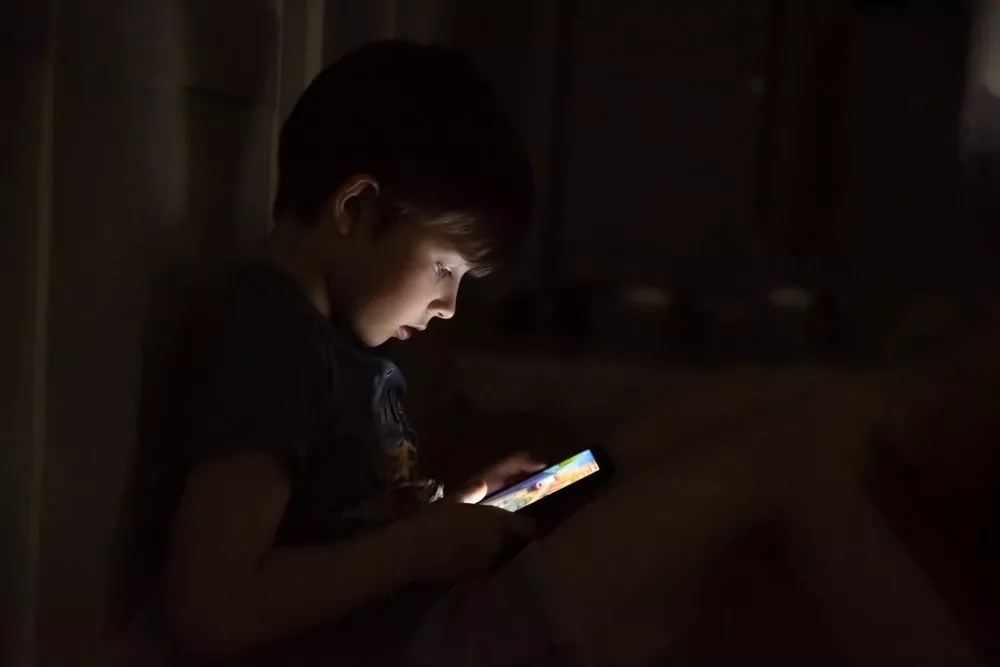 En ung dreng sidder på gulvet i et mørkt rum, oplyst af lyset fra en smartphone-skærm, som han kigger intenst på.