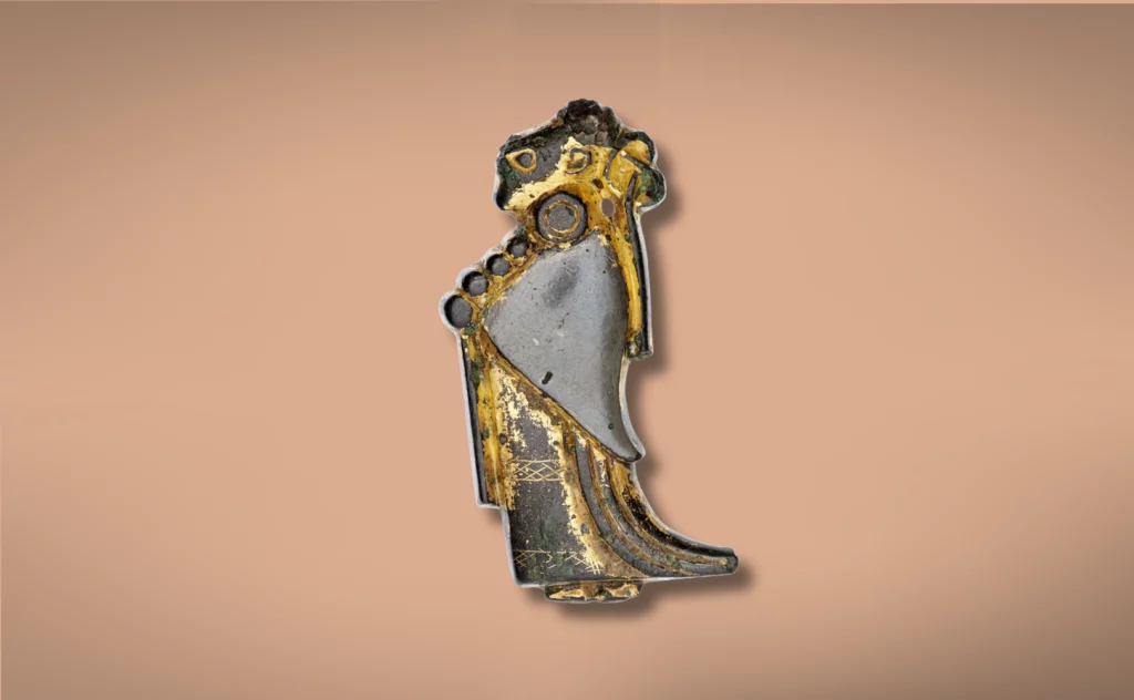 En lille, udsmykket guld- og sølvgenstand formet som en stiliseret menneskefigur i profil med indgraverede detaljer og en buet form på en almindelig beige baggrund.