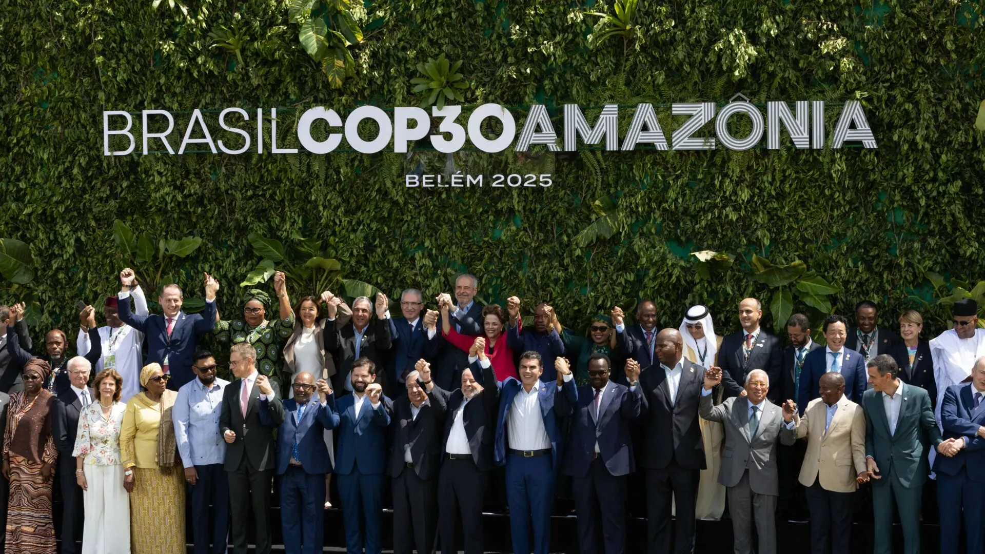 En stor gruppe globale ledere og embedsmænd står sammen udenfor, smilende og med hinanden i hånden, foran en frodig grøn mur med teksten Brasil COP30 Amazônia Belém 2025.