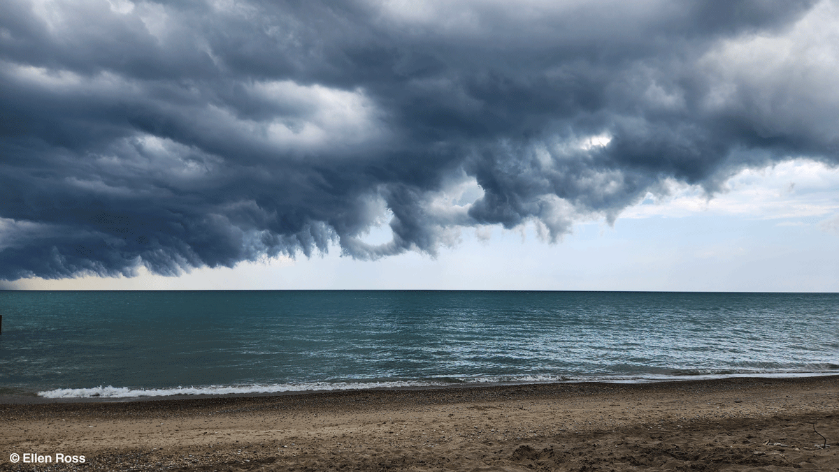 Mørke, dramatiske skyer hænger lavt over en rolig, blå sø med en sandstrand i forgrunden. Himlen virker stormfuld og turbulent og står i kontrast til det stille vand og den tomme kystlinje.