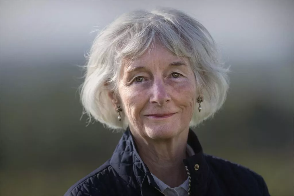 En ældre kvinde med kort gråt hår og lys hud, iført en mørk quiltet jakke og små dinglende øreringe, står udendørs med en naturlig baggrund med blød fokus.
