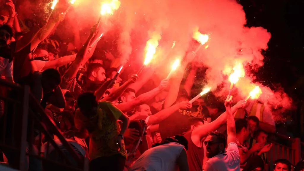 En flok begejstrede fans holder tændte nødblus op, der producerer stærkt rødt lys og røg og skaber en intens og energisk atmosfære ved en sportsbegivenhed.