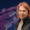 En kvinde med kort, rødblondt hår og læderjakke står foran en sløret baggrund med hænder, der holder en controller til et videospil.