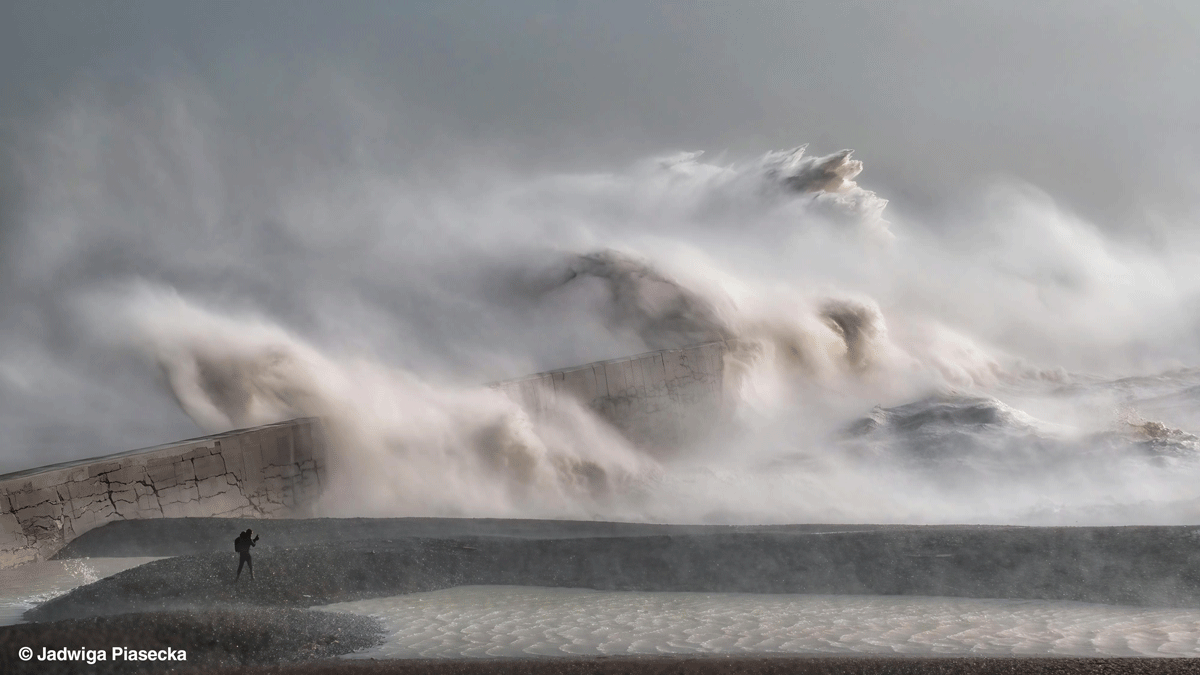 En ensom person står nær en stor, revnet betonmole, mens massive bølger slår ind over den og sender tåge og sprøjt højt op i luften under en overskyet, stormfuld himmel.