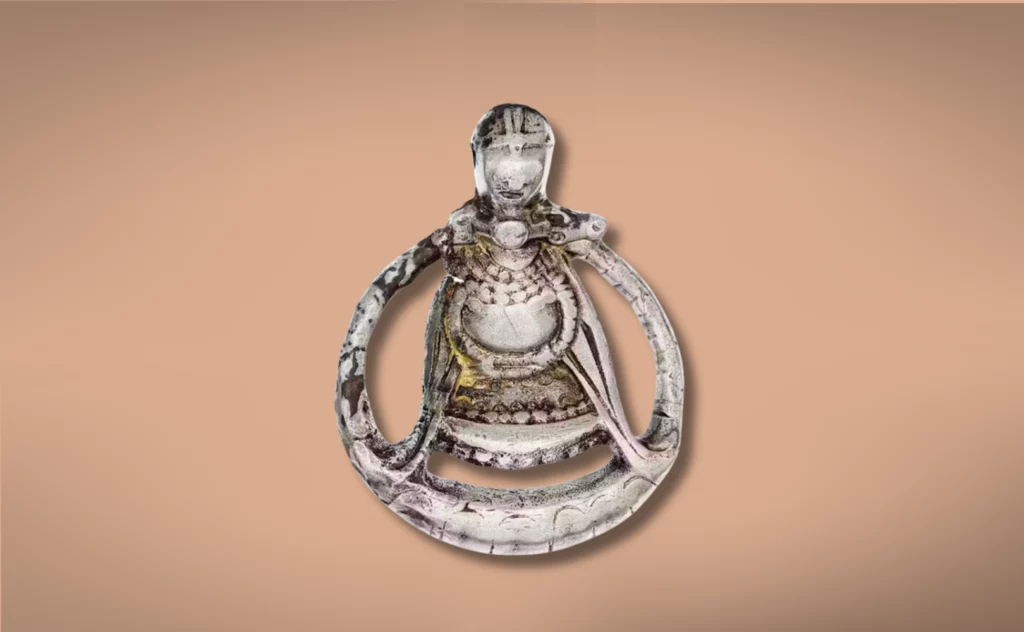 Et metallisk vedhæng fra vikingetiden formet som en kappeklædt figur, med indviklede udskårne detaljer og cirkulær indramning, vist på en almindelig beige baggrund.