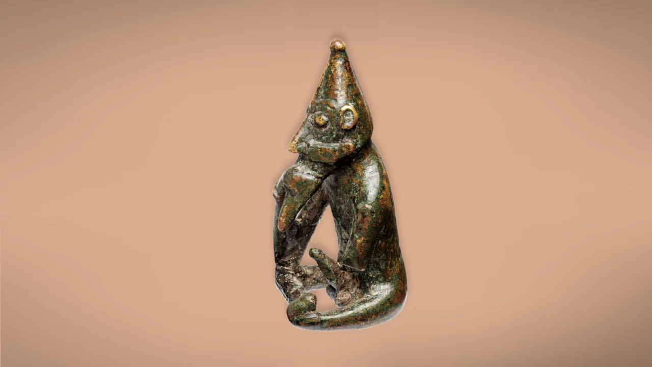 En lille bronzefigur af en siddende abe med spids hat, der hviler hovedet på den ene hånd og har trukket knæene op, på en almindelig beige baggrund.