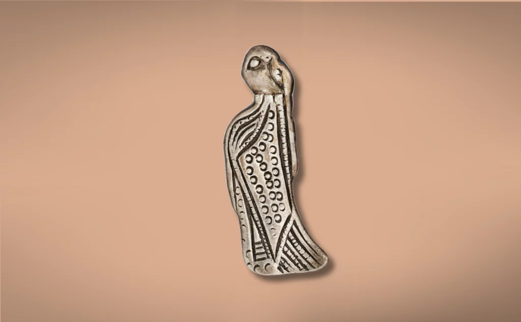 En udskåret, langstrakt figur med geometriske mønstre og prikker på kroppen og et lille, karakterløst hoved. Figuren står på en almindelig lysebrun baggrund.