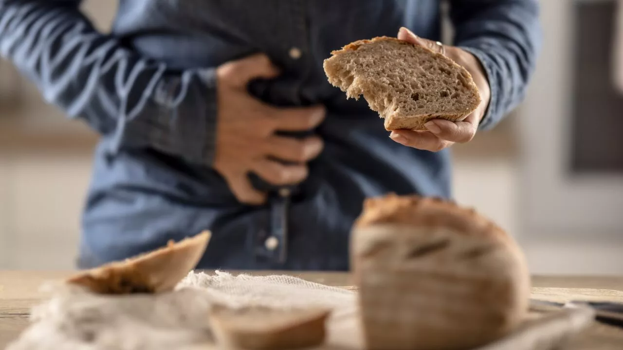 En person i denimskjorte holder sig for maven med den ene hånd og et stykke brød med den anden, hvilket antyder ubehag, med et brød og brødskiver på et bord i forgrunden.