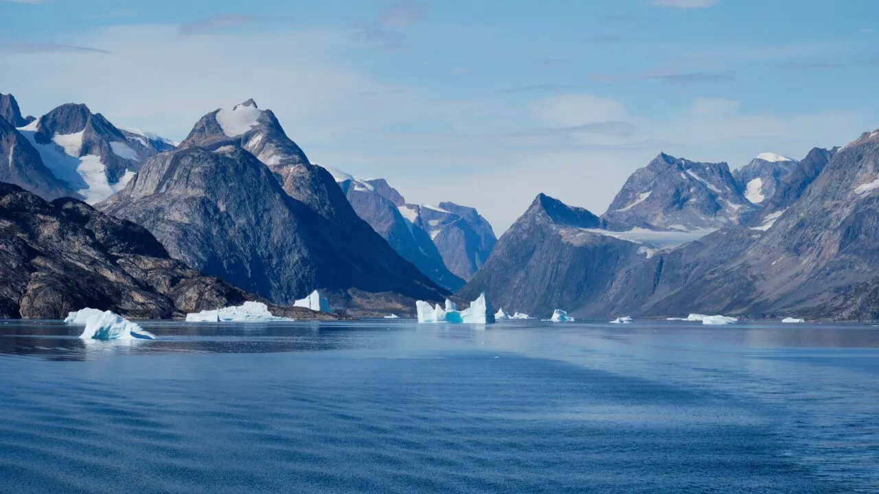 Sneklædte bjerge rejser sig over en rolig, blå fjord med adskillige isbjerge, der flyder på vandet under en klar himmel. Landskabet er barskt og uberørt og giver en fornemmelse af afsidesliggende vildmark.