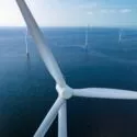 Et nærbillede af en stor hvid vindmølle på havet med flere andre vindmøller synlige i det fjerne over det rolige blå vand under en klar himmel.