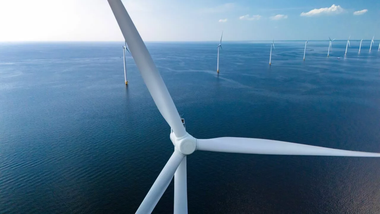 Et nærbillede af en stor hvid vindmølle på havet med flere andre vindmøller synlige i det fjerne over det rolige blå vand under en klar himmel.