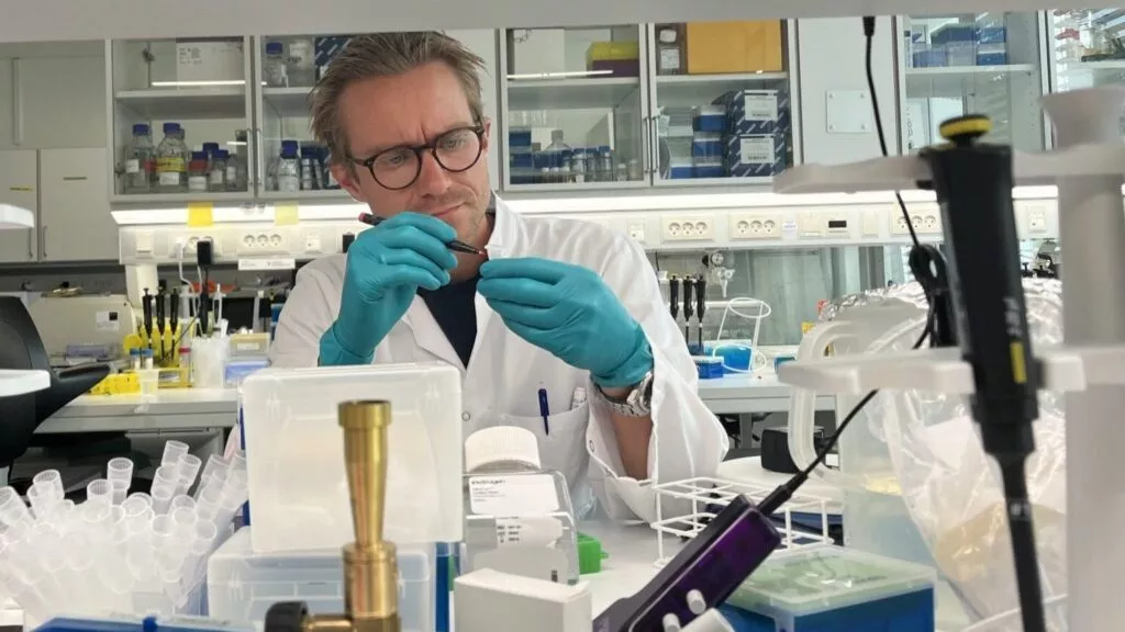 En forsker iført hvid kittel, blågrønne handsker og briller arbejder med laboratorieudstyr ved en rodet laboratoriebænk fyldt med rør, pipetter og beholdere. I baggrunden ses hylder med flasker og forsyninger.