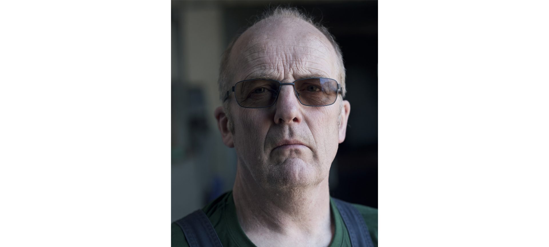 En midaldrende mand med kort hår, iført mørke solbriller, en grøn skjorte og mørke seler, ser direkte ind i kameraet med et alvorligt udtryk. Baggrunden er sløret.