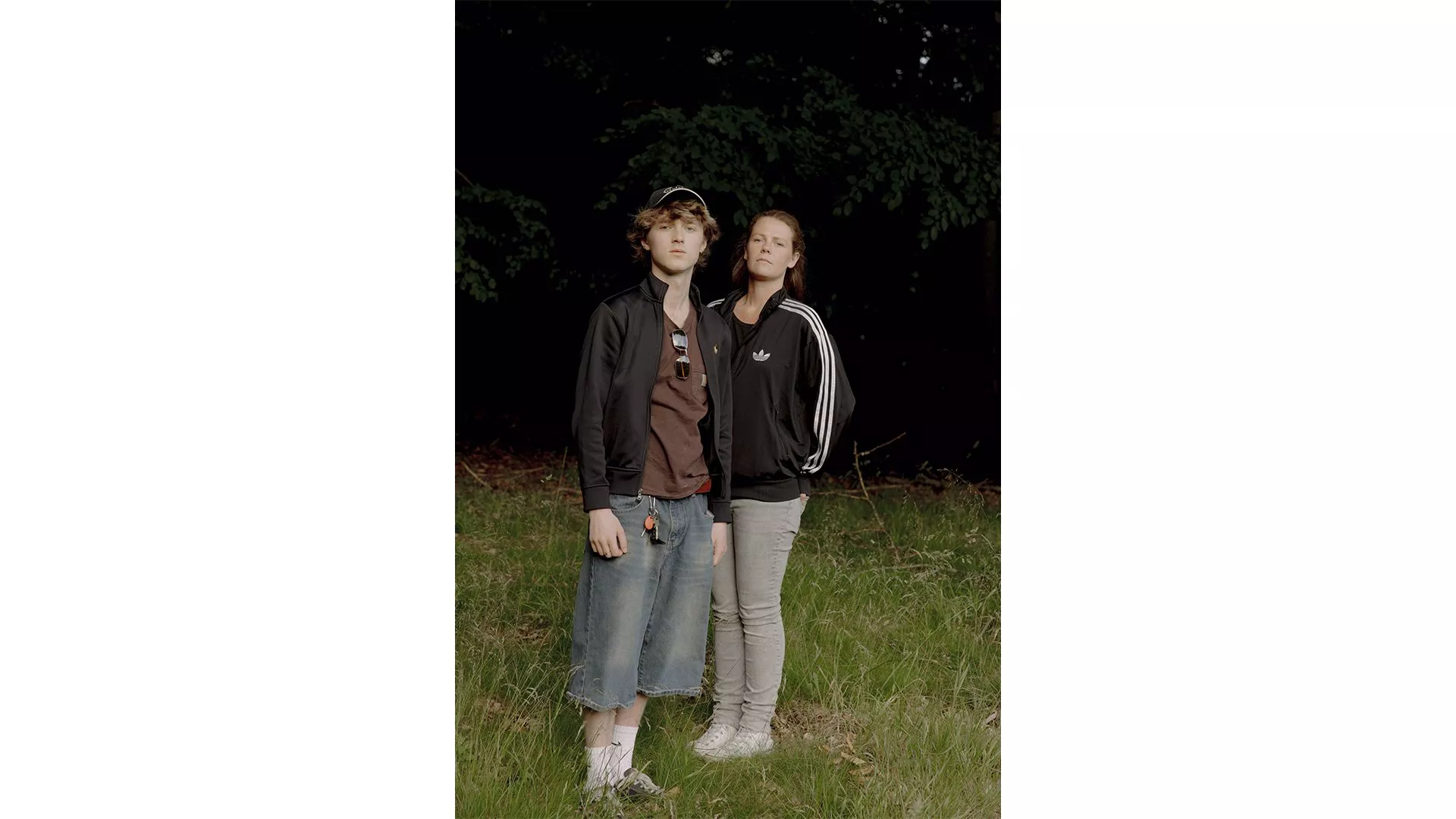 To unge mennesker står udenfor på græsset, begge iført mørke jakker og neutrale ansigtsudtryk. Den ene har kasket, brun skjorte og knælange denimshorts på; den anden har hestehale, grå bukser og sneakers. Træer er synlige i baggrunden.