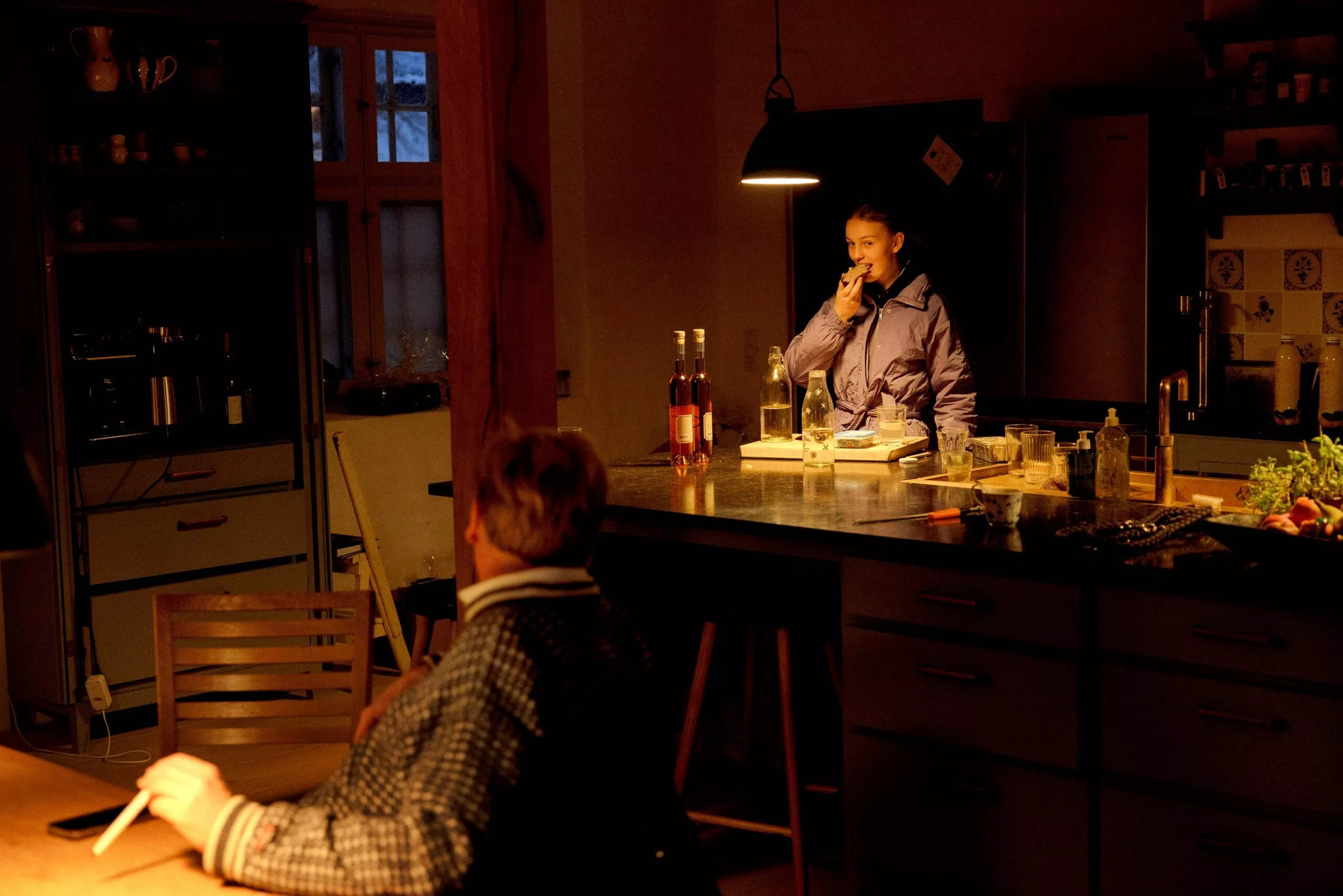 Et svagt oplyst køkken med en person i frakke, der står ved disken, smiler og taler i telefon, mens en anden person sidder ved et bord over for dem. Flasker og køkkenting står på disken.
