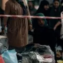 En kvinde med grønt tørklæde sidder blandt poser med ejendele bag afspærringstape, mens andre mennesker står og samler sig i baggrunden, hvilket antyder en scene med fordrivelse eller uddeling af hjælp.