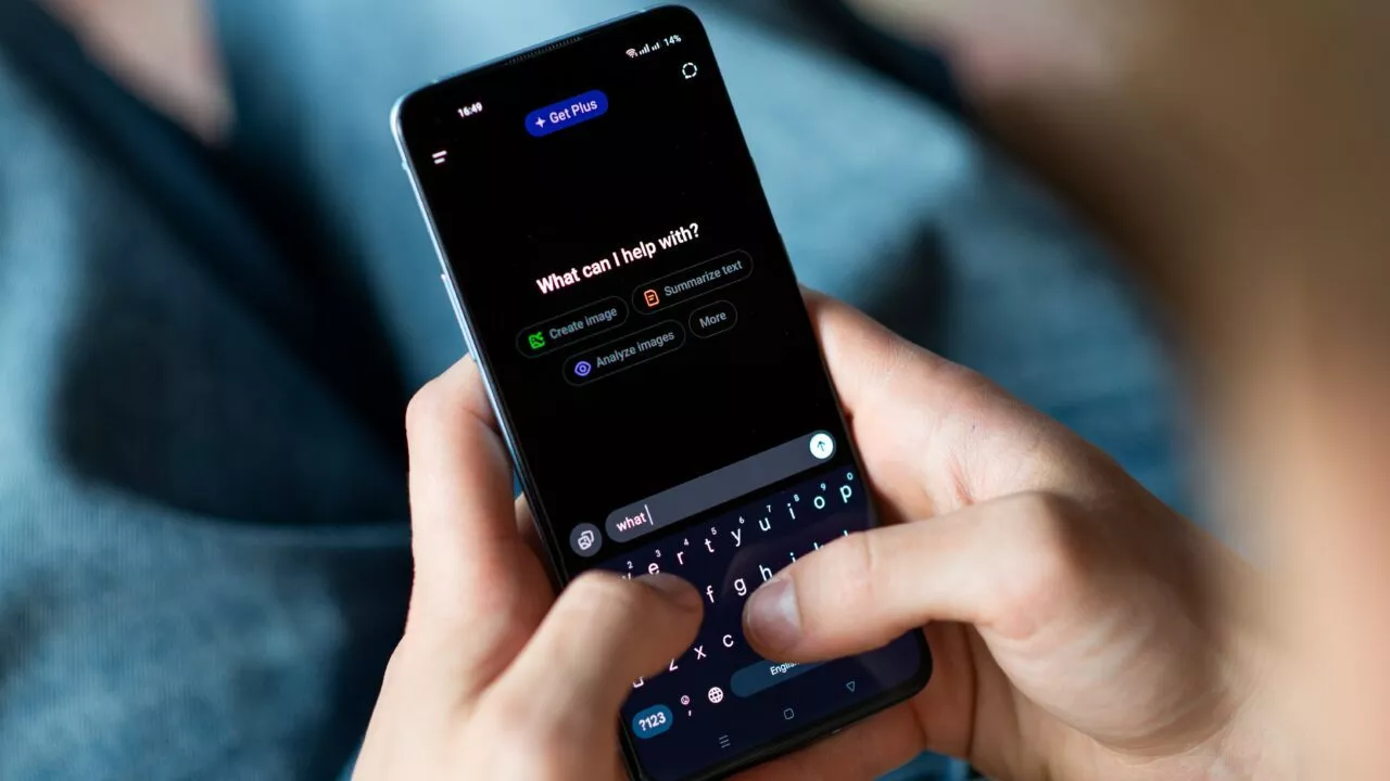 En person holder en smartphone og skriver "what c" i et chatinterface, der spørger: "Hvad kan jeg hjælpe med?" på en mørk skærm med foreslåede prompts vist nedenfor. De sidder med synlige blå jeans.