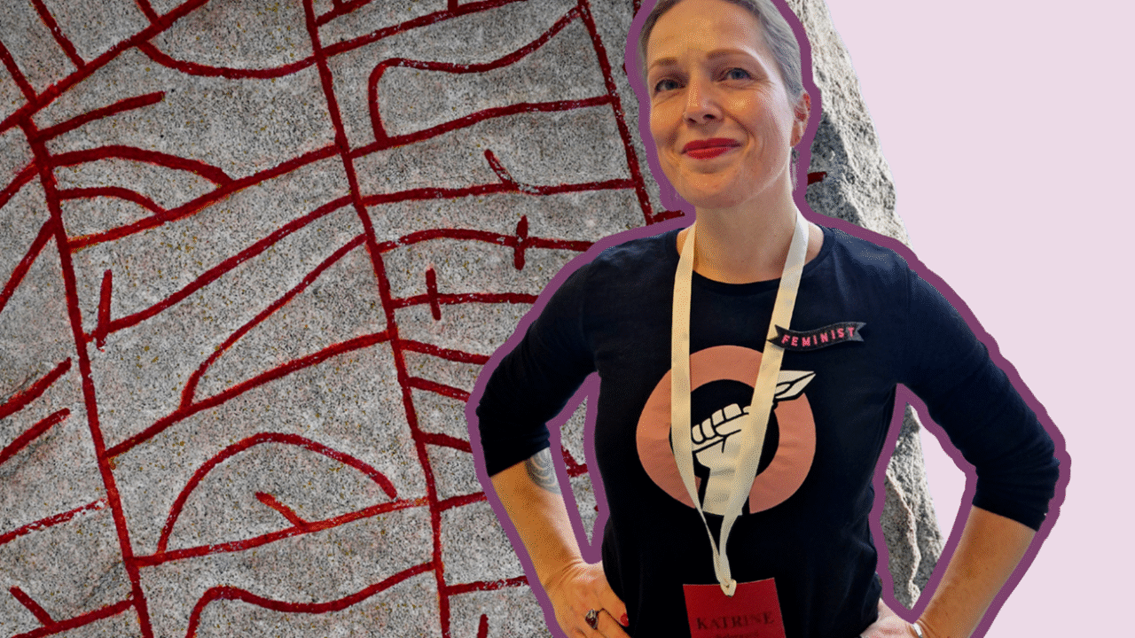 En kvinde står med hænderne på hofterne, iført en sort feministisk t-shirt og navneskilt. Bag hende står en stor sten med røde runer på en lyslilla baggrund.