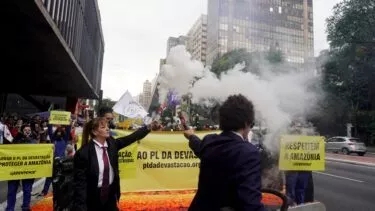 En protest finder sted på en gade i byen. Folk holder et stort gult banner og skilte med krav om respekt for Amazonas. Røg stiger op fra et nødblus, og bygninger står på begge sider af gaden. Nogle demonstranter bærer jakkesæt og skjolde.