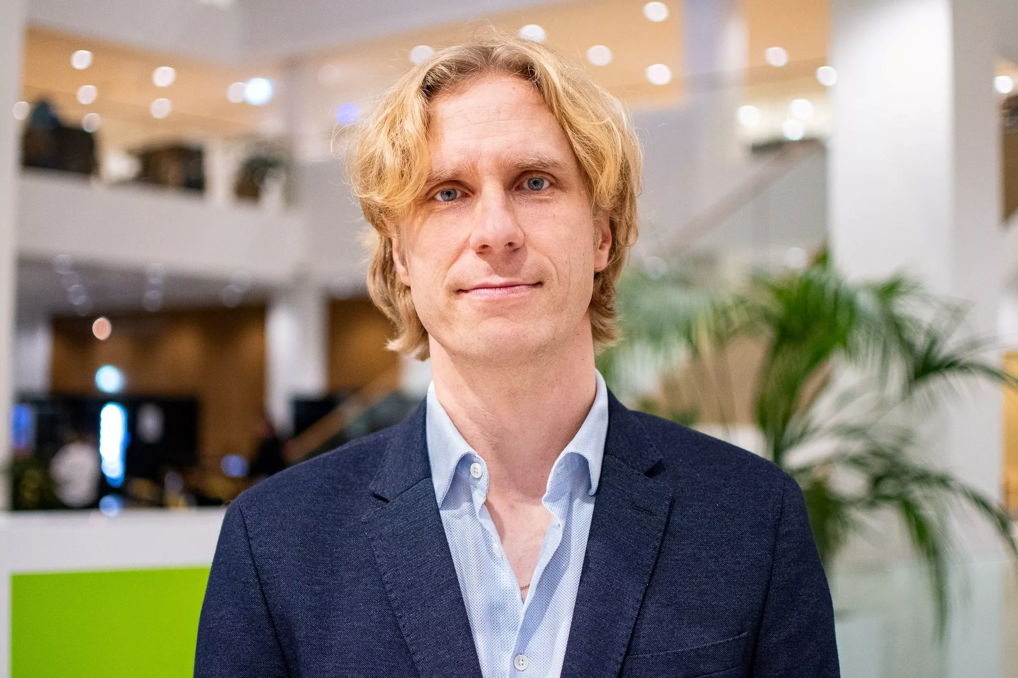 En mand med lyst, bølget hår iført blå habitjakke og lyseblå skjorte står indendørs i et stærkt oplyst moderne kontor med slørede planter og lys i baggrunden.