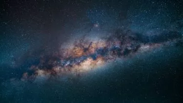 Et levende billede af Mælkevejen strækker sig over en mørk nattehimmel og viser tætte klynger af stjerner, glødende støvskyer og dybe kosmiske farver fra blå til orange.