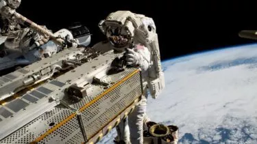 En astronaut i rumdragt arbejder på ydersiden af et rumfartøj med Jorden og dens skyer synlige i baggrunden mod rummets mørke.