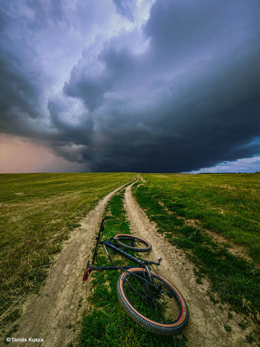En cykel ligger på en grussti, der snor sig gennem en græsmark under en dramatisk, mørk stormhimmel med tykke skyer, der nærmer sig.