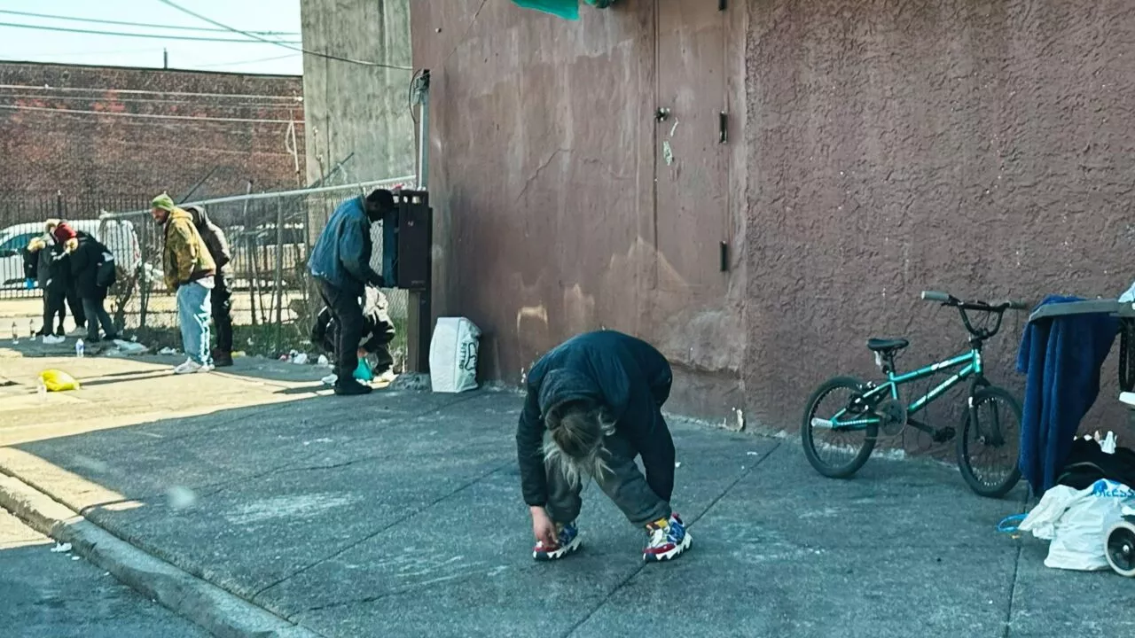 En person bøjer sig forover på et fortov nær en mur, mens andre står og sidder i nærheden. En cykel ligger på jorden til højre, og forskellige ejendele er spredt rundt omkring. Scenen virker urban og dyster.