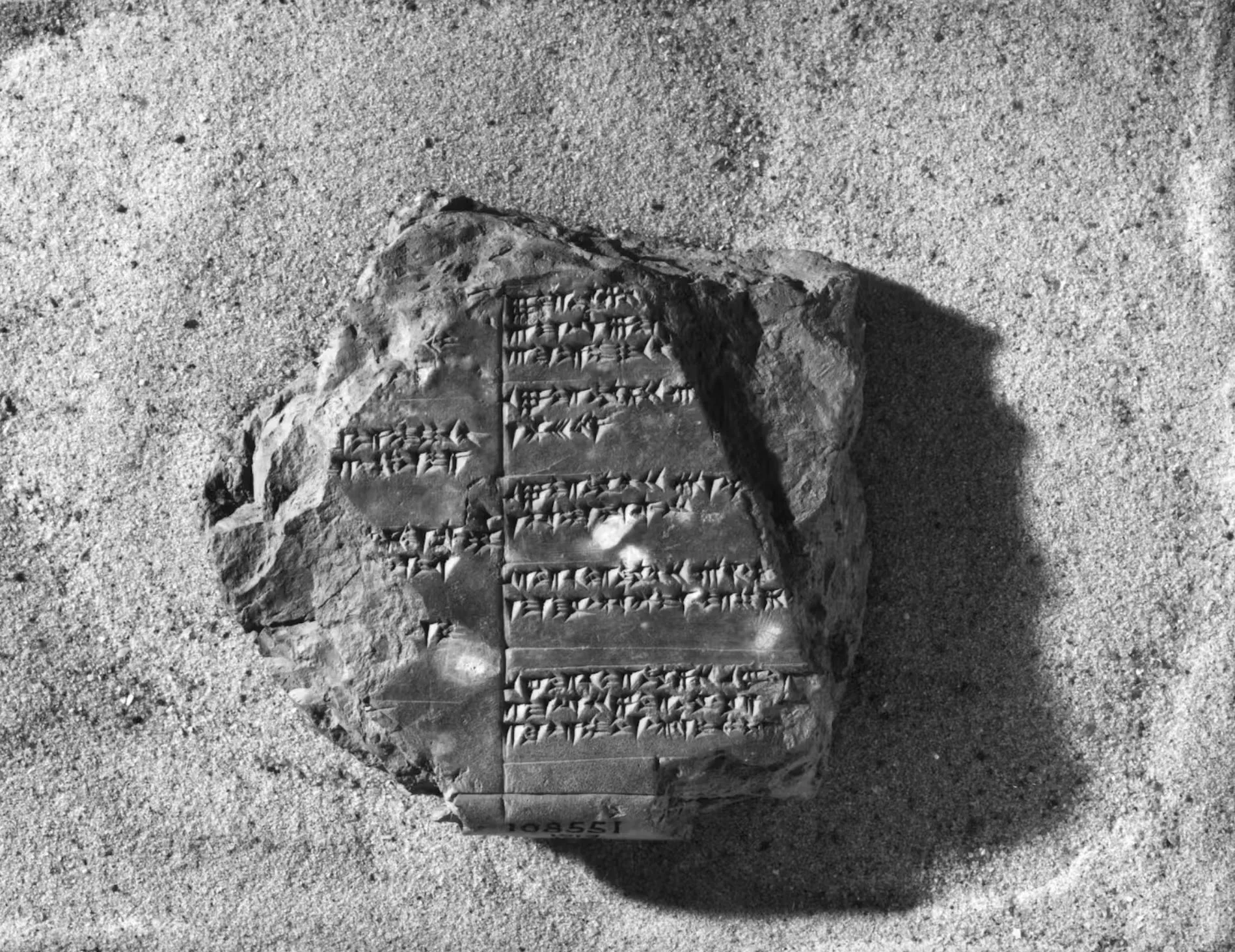 En stentavle med gamle kileskriftinskriptioner hviler på en sandet overflade. De indgraverede symboler er arrangeret i pæne vandrette linjer på tværs af den mørke, uregelmæssigt formede tavle.
