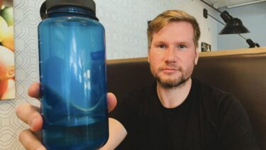 En mand med kort lysebrunt hår og skæg, iført sort t-shirt, holder en blå plastikvandflaske med sort låg op, mens han sidder på en café med mønstret tapet og vægkunst - muligvis kemikeren Alexander Sandtorv.