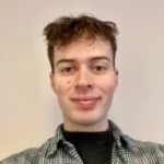 En ung person med kort, bølget brunt hår smiler til kameraet. Vedkommende har en sort skjorte på under en ternet skjorte med knapper. Baggrunden er ensfarvet og lys.
