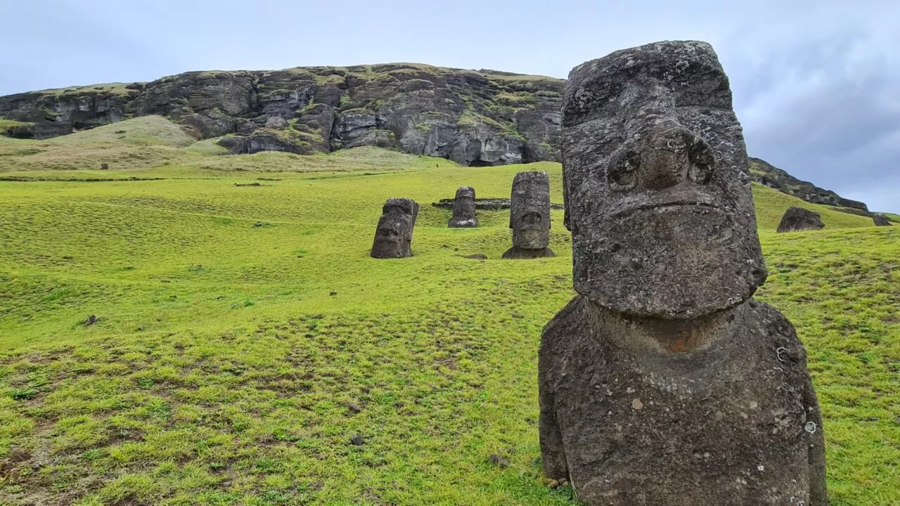 Moai-statuer står på en græsklædt bjergskråning med en klippeklint i baggrunden under en overskyet himmel på Påskeøen.