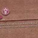 En murstensvæg med Københavns Universitets segl og navn på dansk og engelsk, med et rødbrunt cirkulært emblem og enkle geometriske mønstre over teksten.