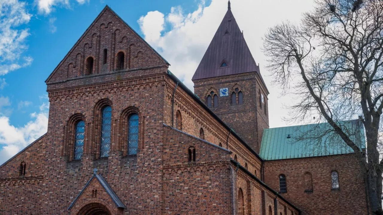 En stor murstenskirke med buede vinduer og et højt klokketårn står mod en delvis overskyet himmel. Nøgne træer indrammer bygningens højre side.