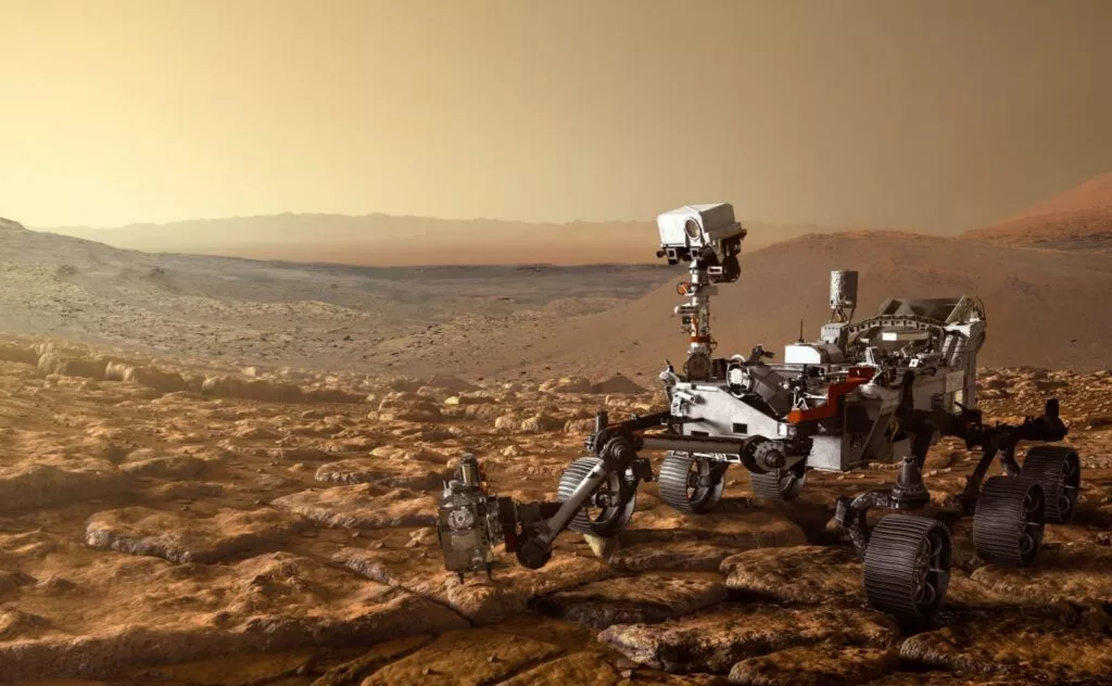 En Mars-robot med seks hjul udforsker et stenet, rødbrunt Mars-landskab under en diset, orange himmel med fjerne bjerge i baggrunden.