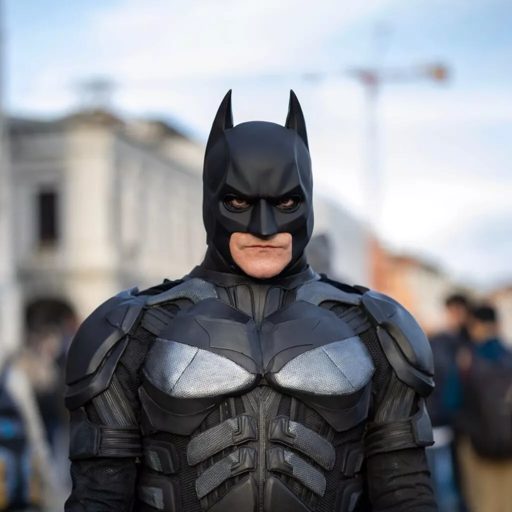 En person klædt i et detaljeret Batman-kostume står udendørs med bygninger og mennesker sløret i baggrunden. Kostumet er sort med pansrede detaljer, og personen bærer den ikoniske flagermusmaske.