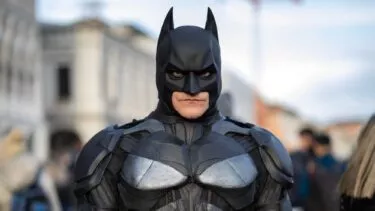 En person klædt i et detaljeret Batman-kostume står udendørs med bygninger og mennesker sløret i baggrunden. Kostumet er sort med pansrede detaljer, og personen bærer den ikoniske flagermusmaske.