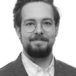 En mand med briller, overskæg og skæg, iført habitjakke og hvid skjorte med krave, smiler let, mens han kigger ind i kameraet. Billedet er i sort-hvid.