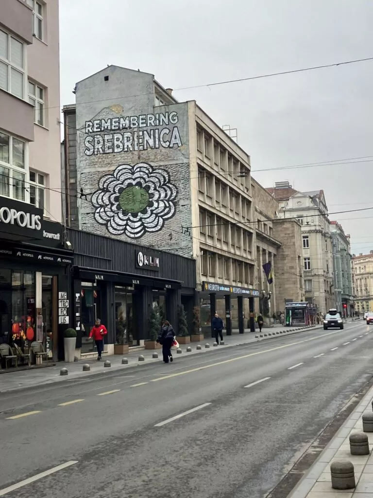 En bygade med et vægmaleri på en bygning, hvor der står Remembering Srebrenica ved siden af et hvidt blomsterdesign. Folk går på fortovet under en overskyet himmel, og flere butikker ligger langs gaden.