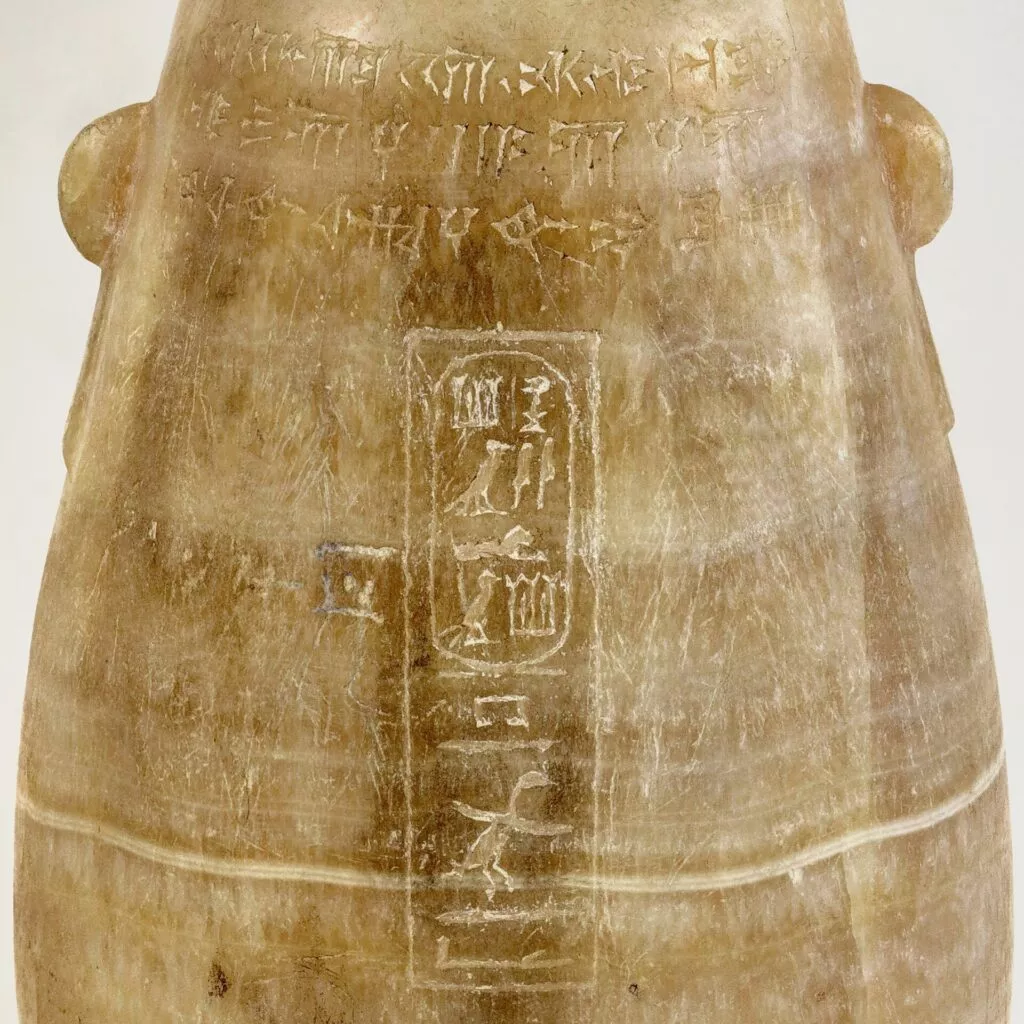 Nærbillede af en gammel stenartefakt med falmede udskæringer af hieroglyffer og kileskriftsindskrifter. Overfladen er glat og lysebrun og bærer præg af alder.